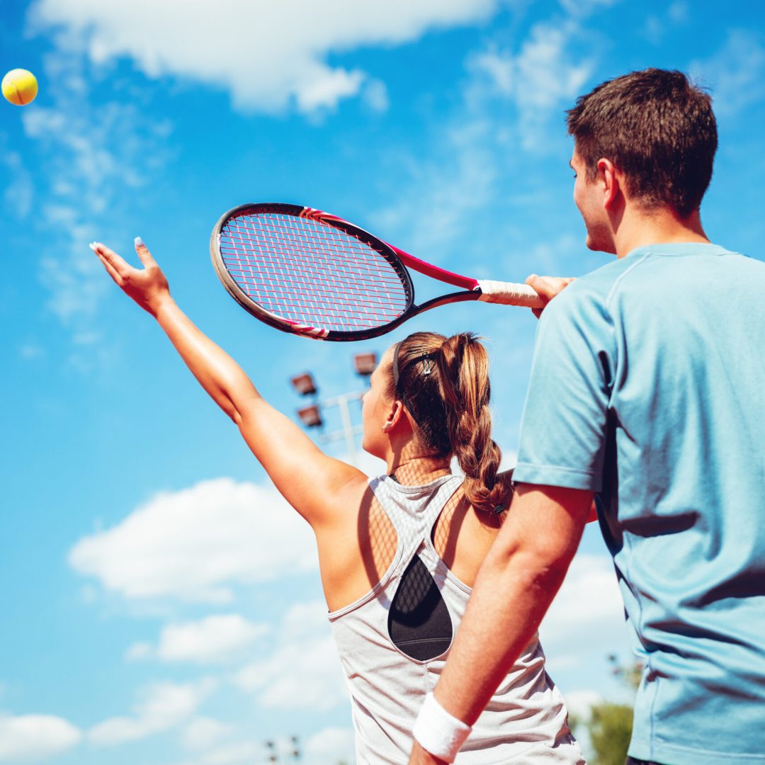 Beautiful young left-handed woman with her trainer practicing serve on outdoor tennis court.